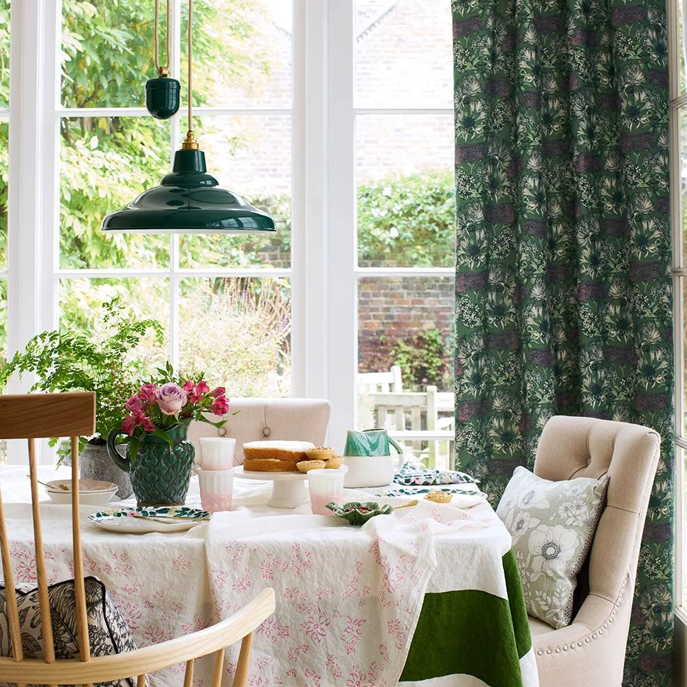 Dark green botanical curtain beside a large pale-framed window and green hanging light, with a dressed dining table and pale upholstered chairs.