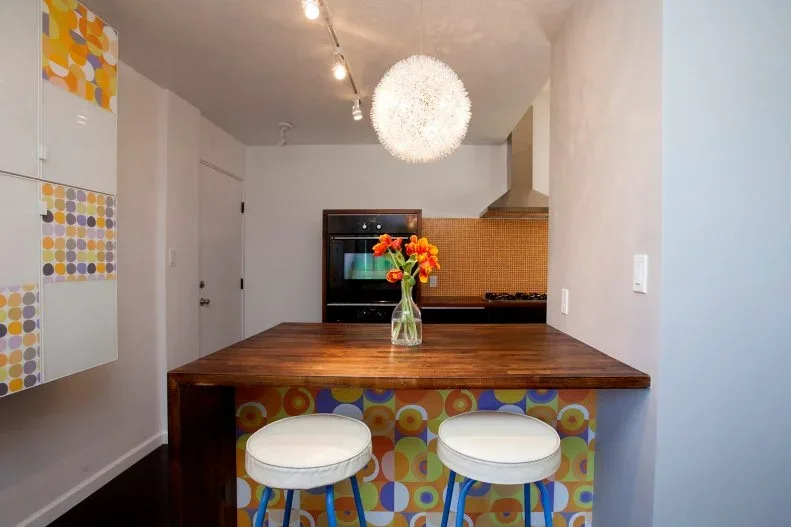 Wood-topped breakfast bar with a multicolored circular pattern, two white stools and pale kitchen walls.