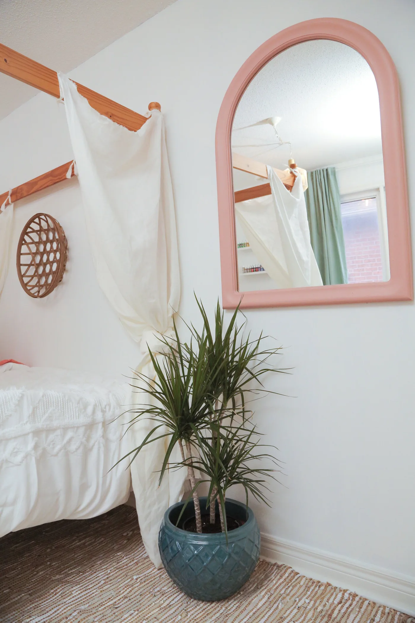 Pink arched wall mirror above a tall green plant in a blue pot beside white bed fabric.