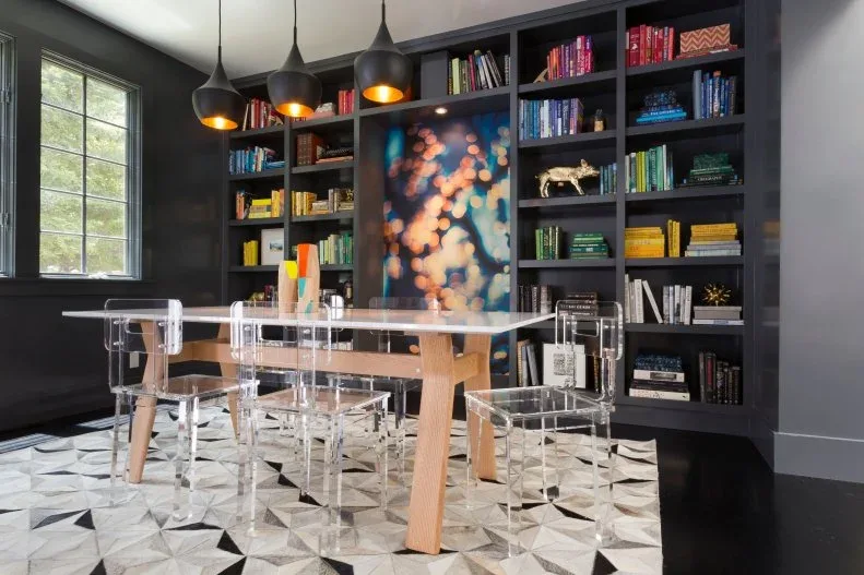 Transparent dining armchairs around a pale wooden table in a black room with colorful bookshelves, dark pendant lights, and a patterned pale floor.
