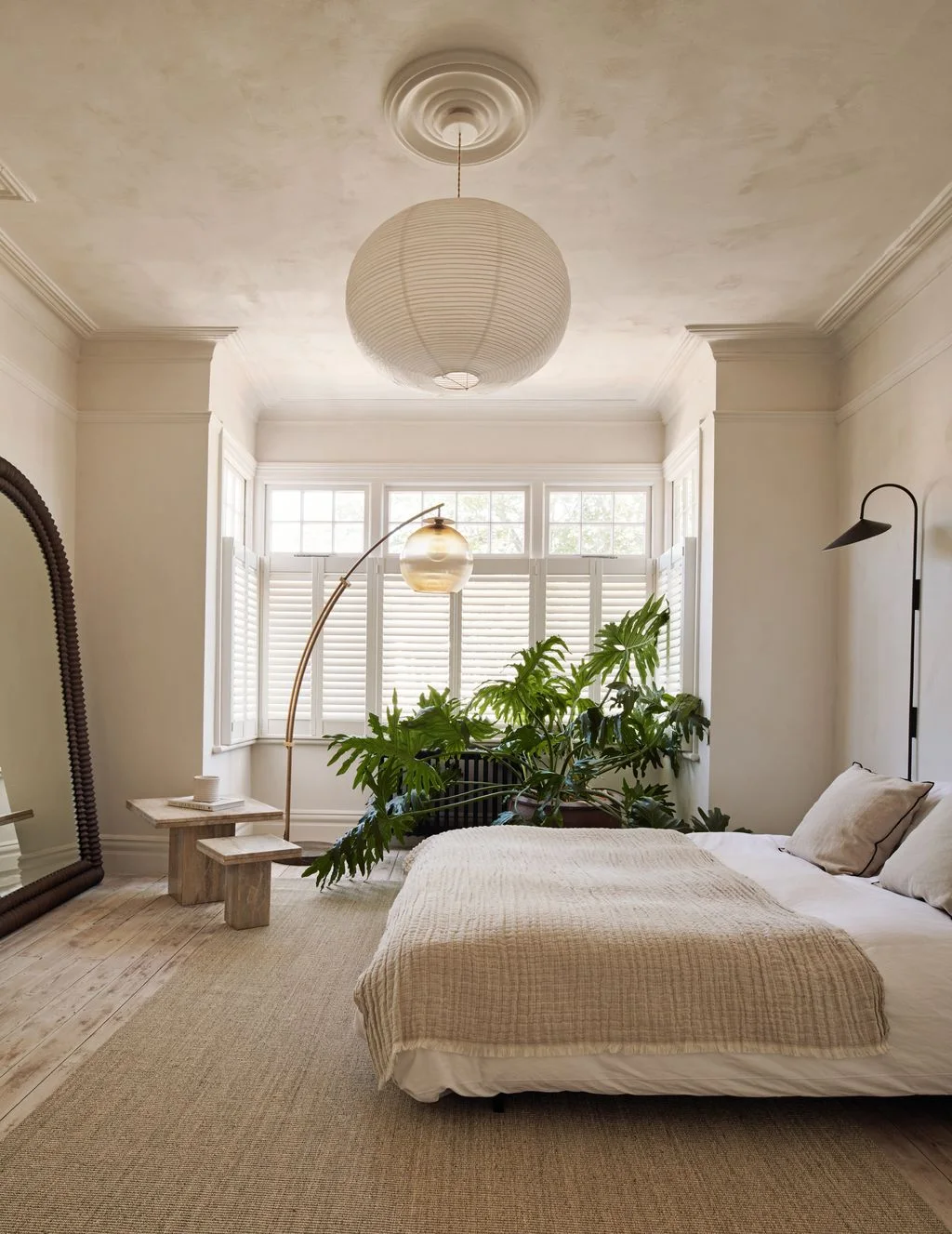 A pale bed stands beside simple wood tables, a curved floor lamp and dark wall lamp beneath a large light pendant near a shuttered bay window