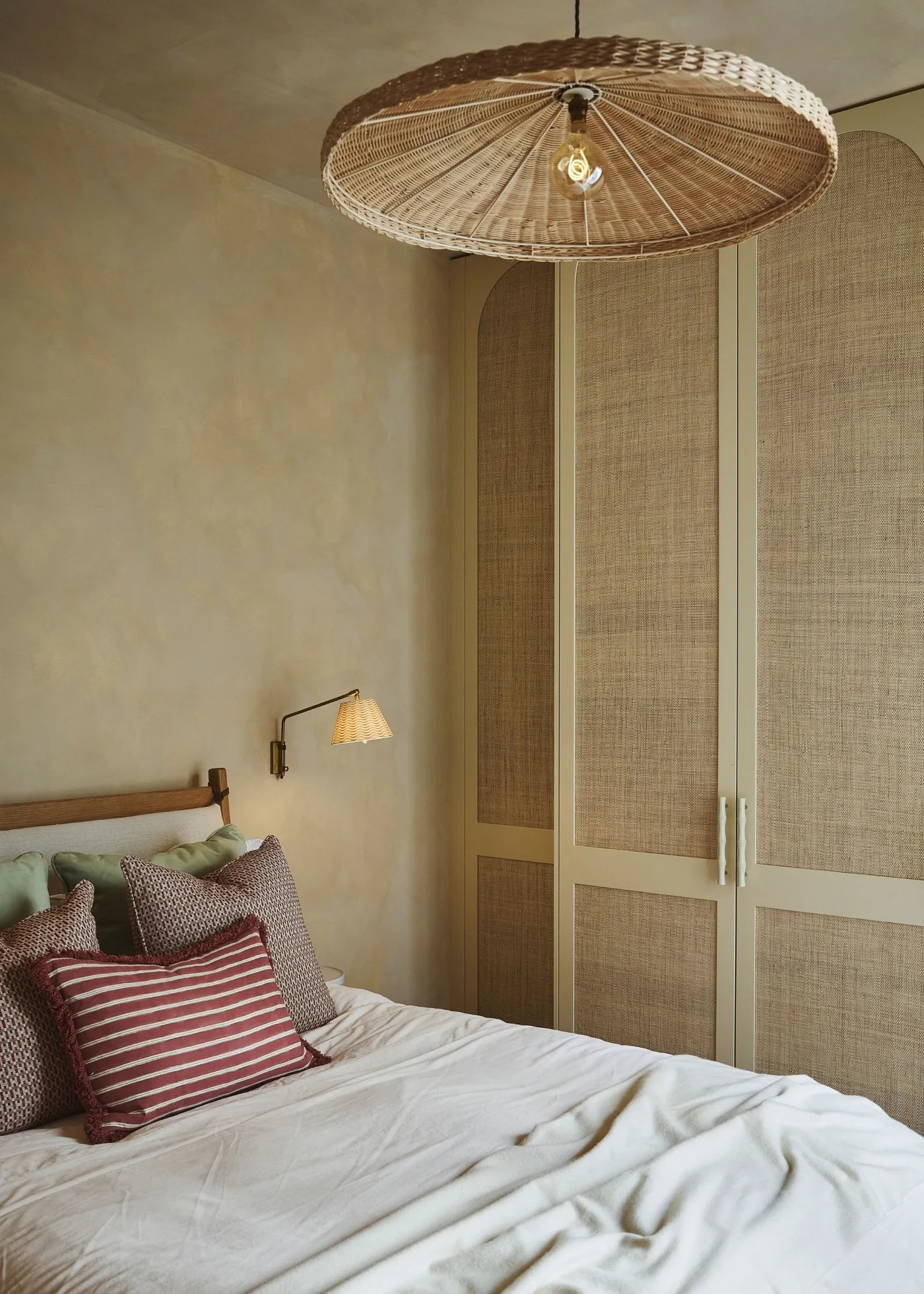 Sand-colored bedroom with mottled walls, woven lighting, tall textured cupboards and bedding in muted rose and green with a rust striped cushion