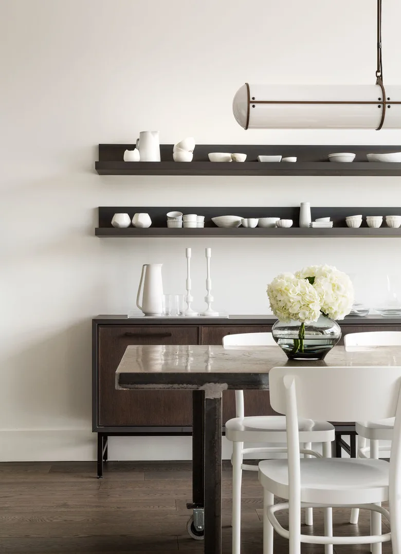 Two dark shelf ledges displaying pale bowls above a dark sideboard behind a dining table with white chairs