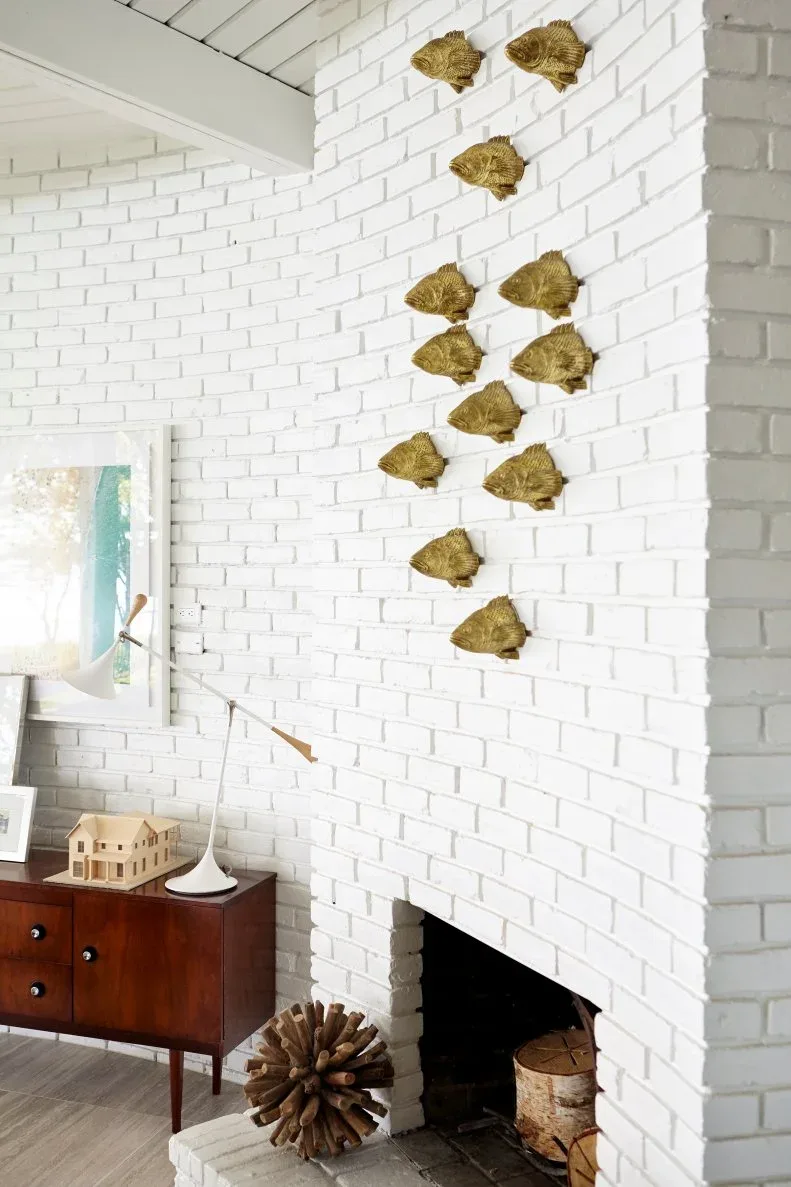 Tall white brick fireplace face with grouped gold-toned fish ornaments beside wood sideboard