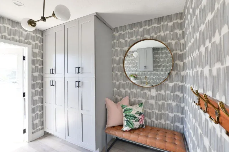 Mudroom with gray patterned walls, pale cupboard, tan tufted bench, round mirror, wooden hook rail and floral cushion.