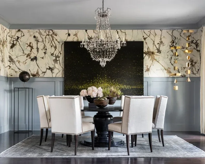 Dark round pedestal table with pale chairs, a chandelier, patterned wall panels and black-and-gold artwork.