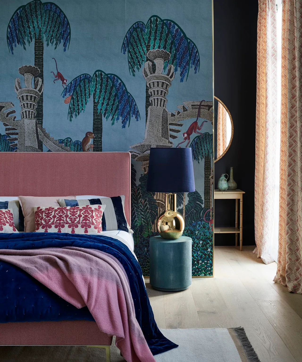 Bedroom with blue tree-and-animal mural, pink headboard, blue bedding, teal bedside table, dark blue lamp and patterned curtain.