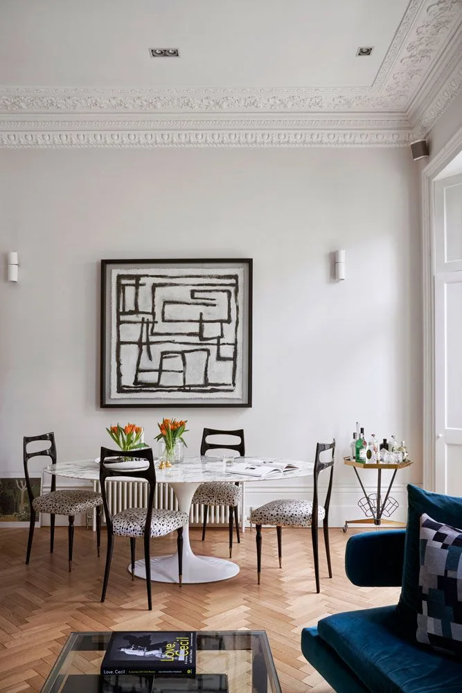 A pale oval pedestal dining table stands among dark open-back chairs with patterned seats beside geometric artwork, a blue sofa and a small drinks table