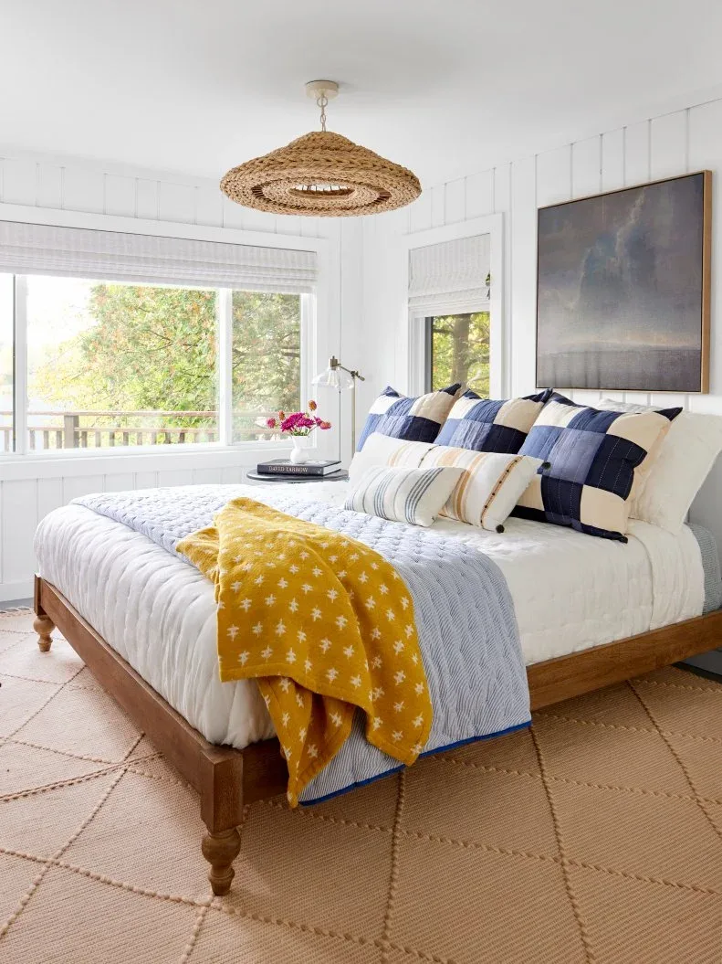 White boarded bedroom with wood bed, blue checked pillows, yellow throw and woven pendant