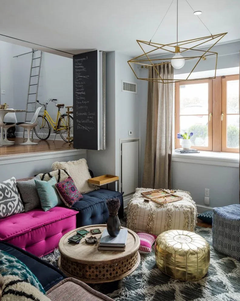 Compact living room with bright pink and dark blue low modular seats, mixed cushions, round patterned table, pale and gold poufs and gray walls.