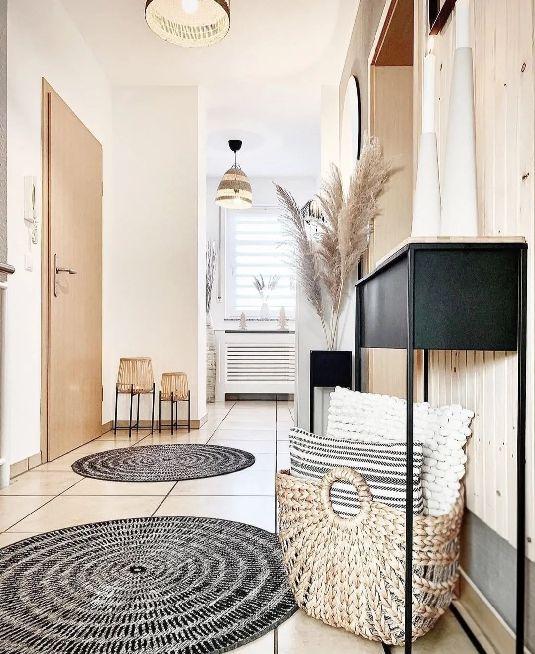 A narrow dark cabinet stands against a pale wooden wall beside a wooden door, woven basket, round patterned rugs and textured pendants