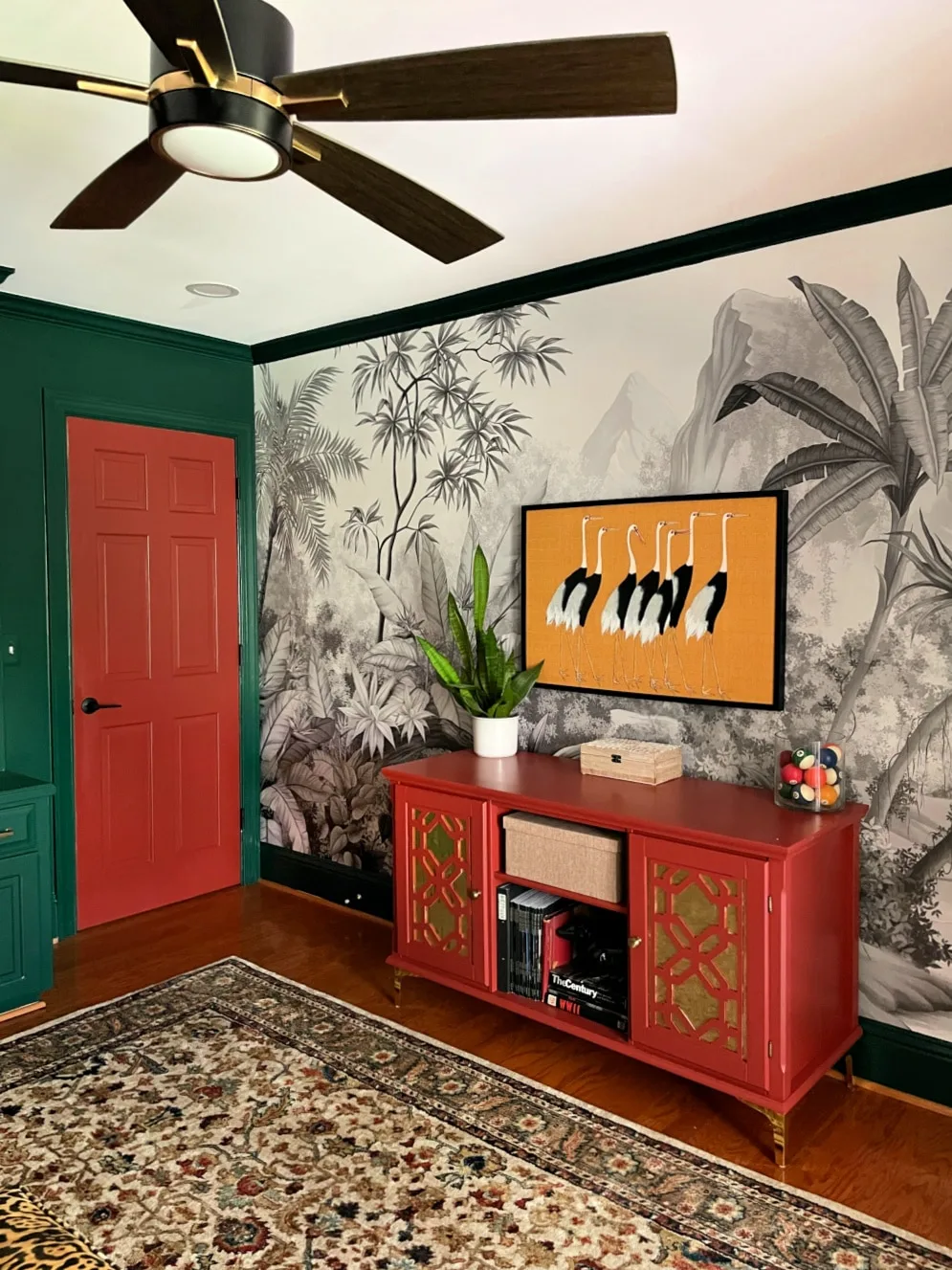 Deep green walls and trim sit beside a red storage cabinet beneath botanical wallpaper and a dark ceiling fan
