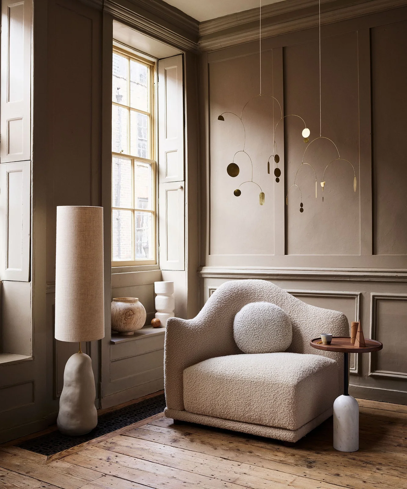 A pale rounded chair with a round cushion stands beside a small wood side table and tall lamp against paneled walls near a window