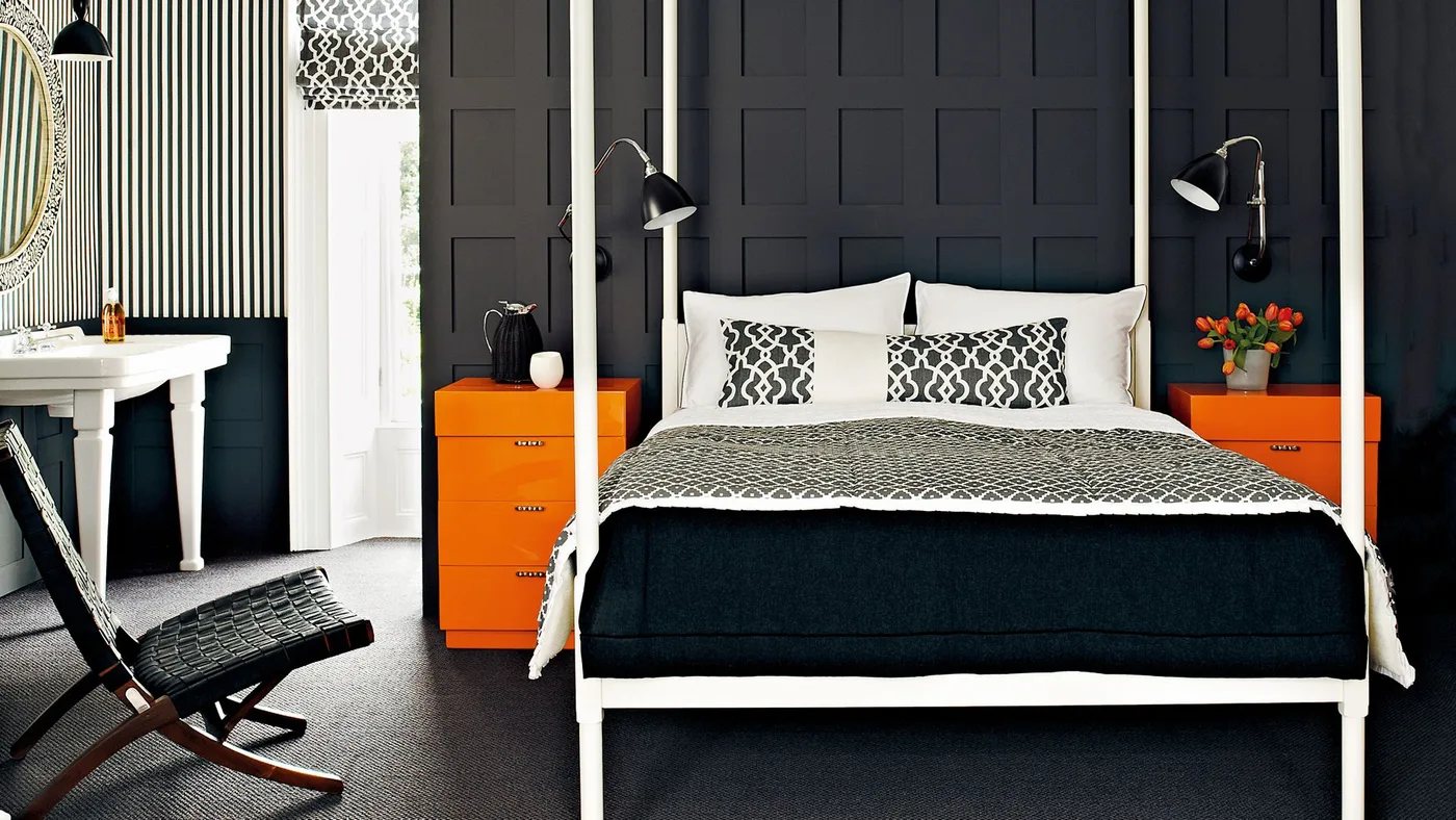 A white four-poster bed stands against charcoal paneling between orange drawer chests with patterned black-and-white bedding