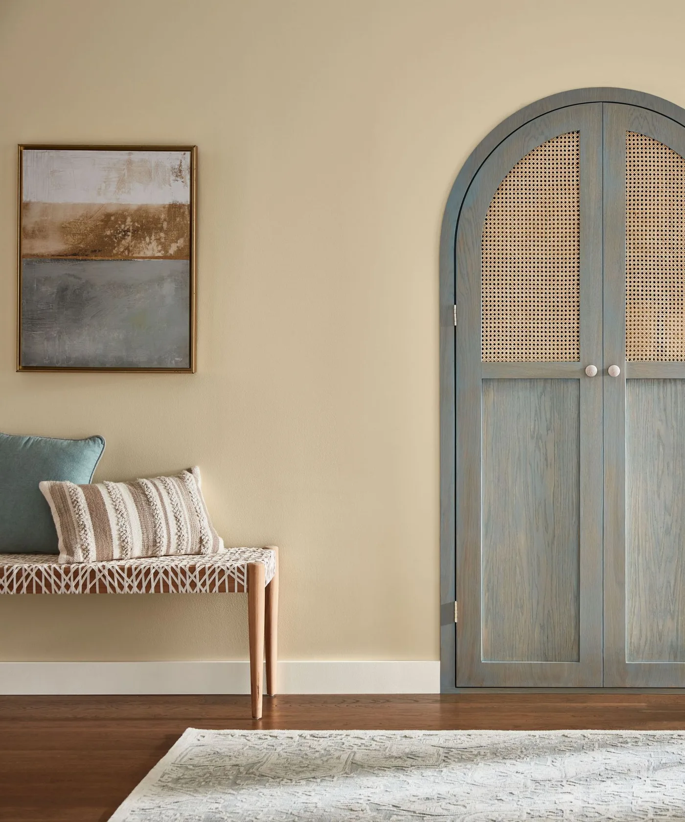 Entrance with tall arched blue-gray woven-panel cupboard, wooden bench, striped cushion, pale wall and framed artwork.