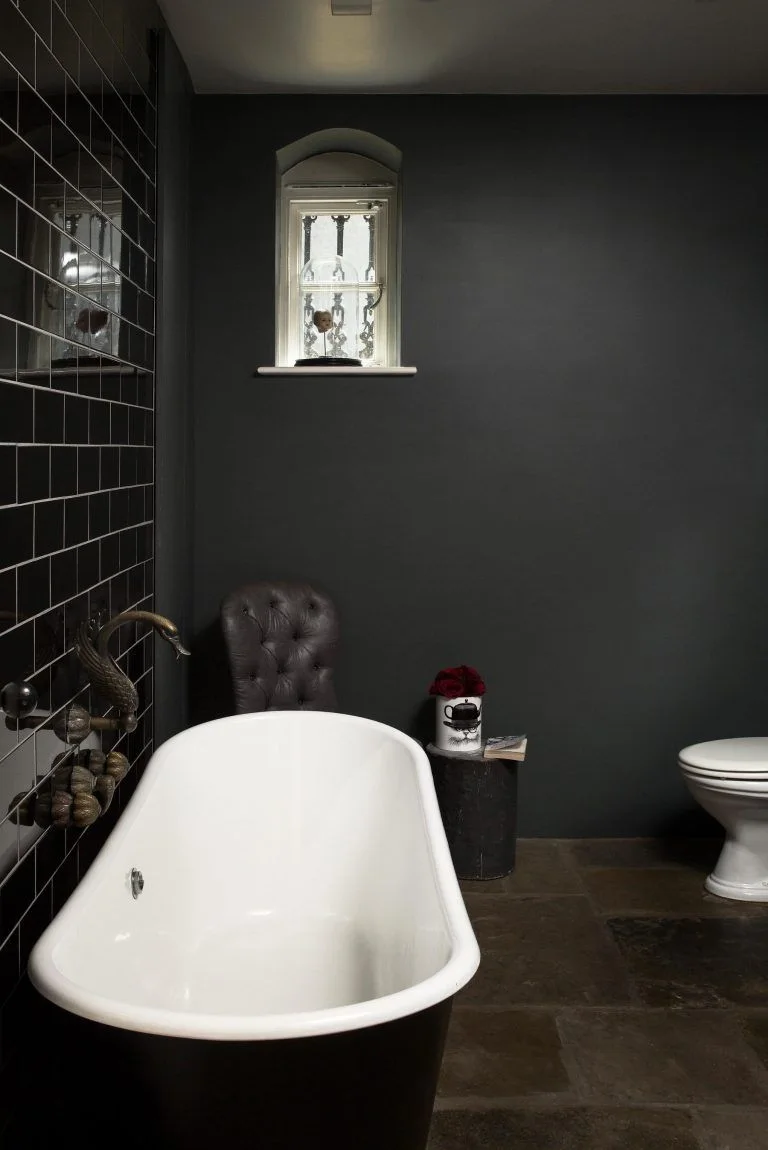 A dark freestanding bathtub with a white interior stands beside black wall tiles, a charcoal painted wall, a narrow window and a white toilet