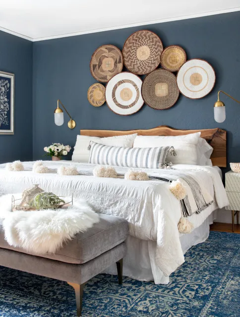 Woven round wall pieces form a cluster above a pale bed against deep blue paint.