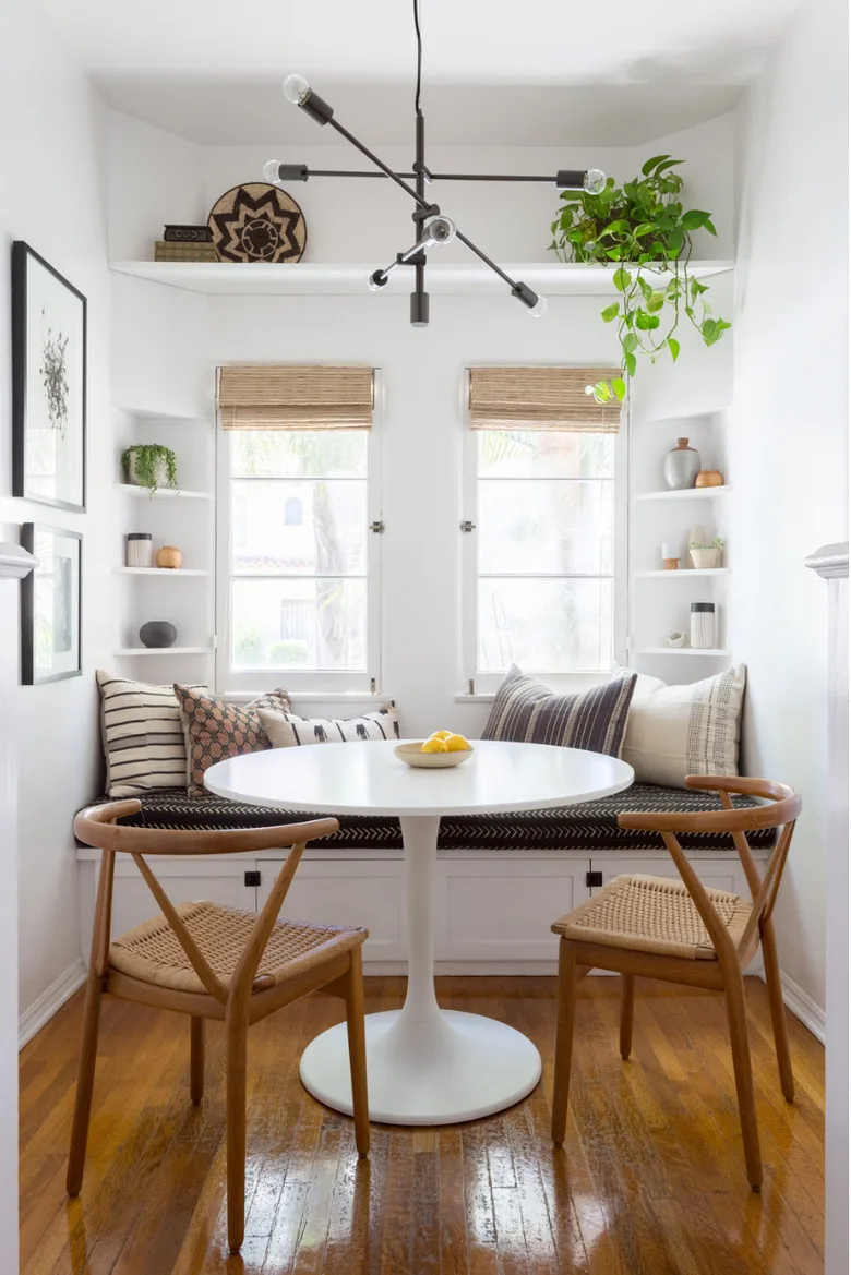 White corner shelves and a high shelf above two windows in a dining nook with a round table and cushioned bench