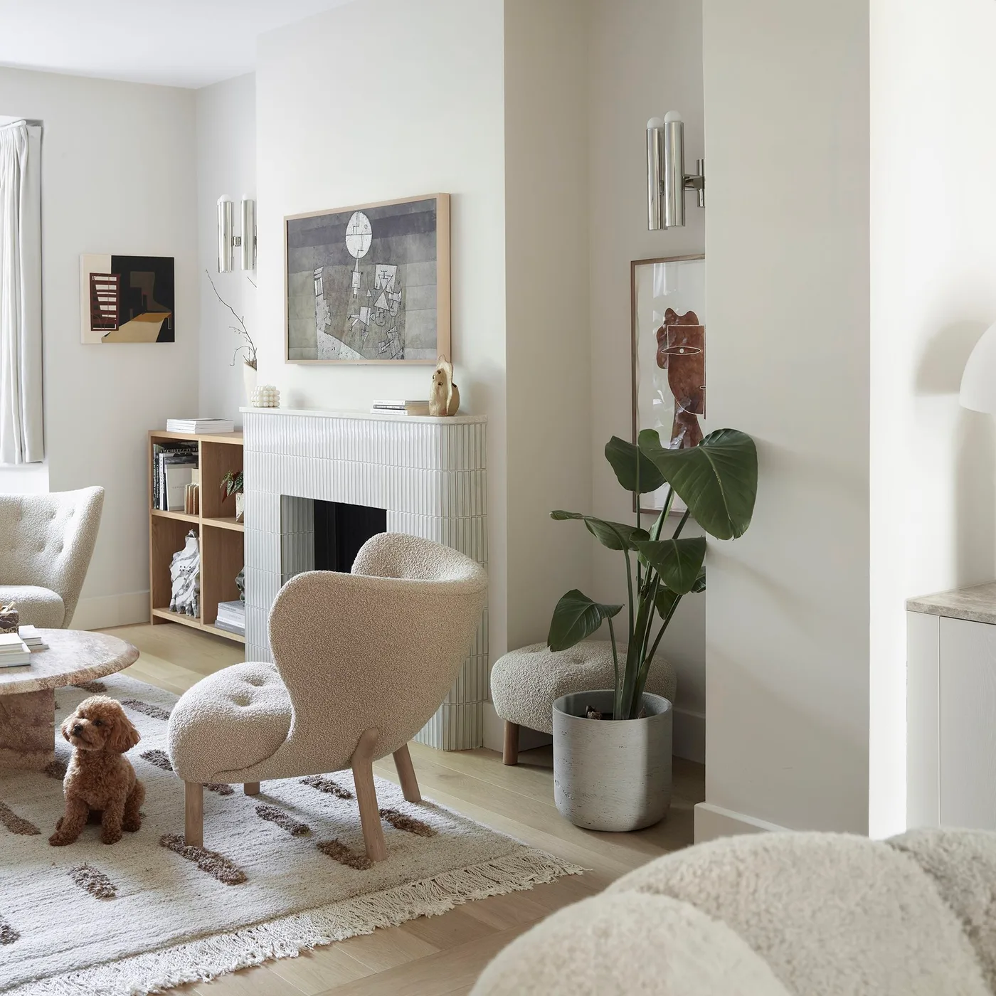 A pale textured armchair stands beside a light fireplace, simple wood shelving, a leafy plant and a pale rug with darker markings