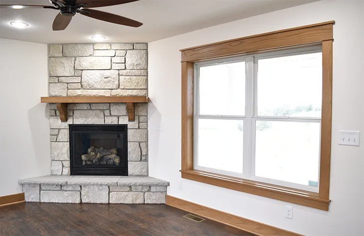 Pale block corner fireplace with a raised hearth and wood mantel beside a wide wood-trimmed window.