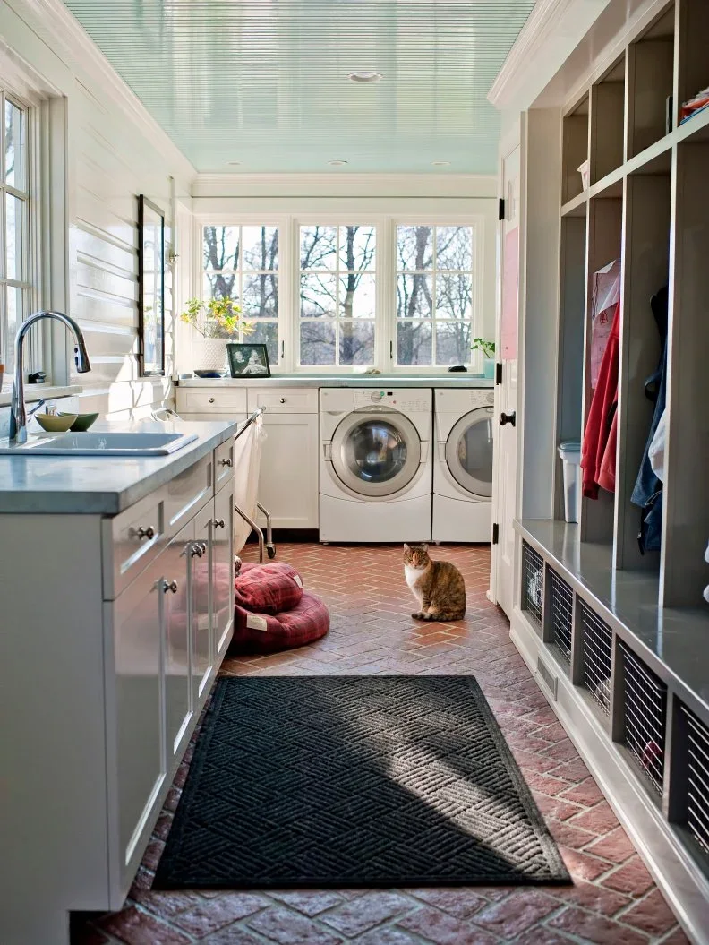 A narrow paneled room contains pale washing appliances and sink storage opposite open coat compartments, with a pale blue ceiling and brick-patterned floor