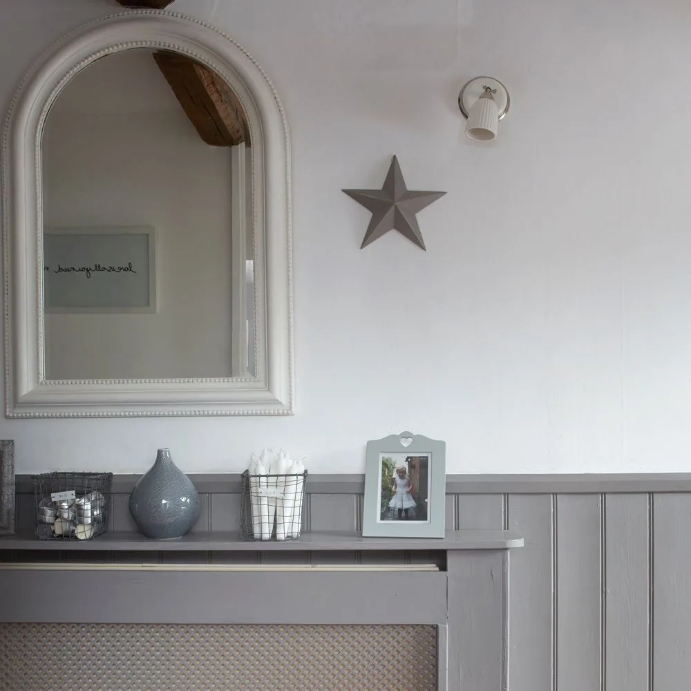 Pale entryway with lower gray paneling, a covered radiator and a pale arched mirror above small decorative objects.