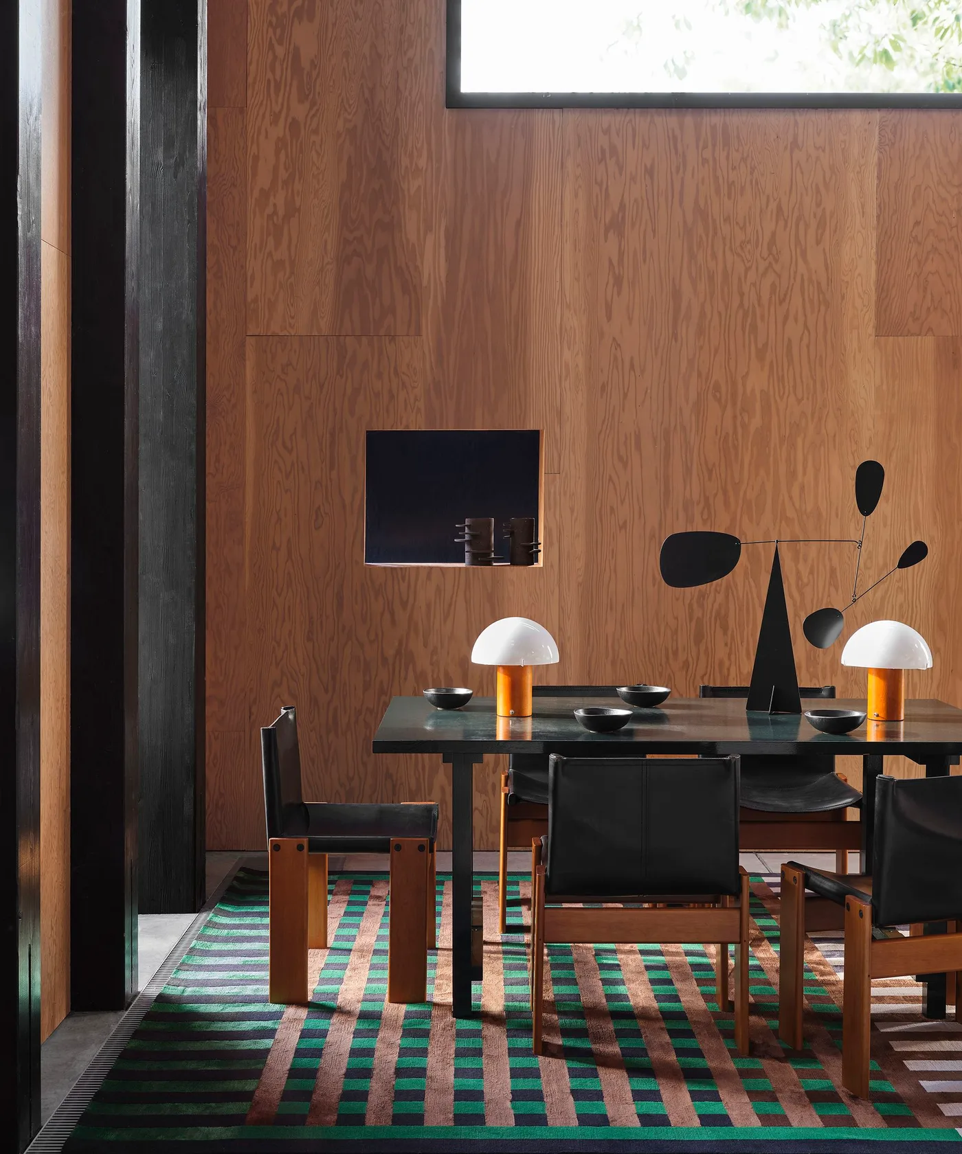 Dining room with warm wood walls, a black table, black seats with wood frames, striped green-pink rug and two small dome lamps