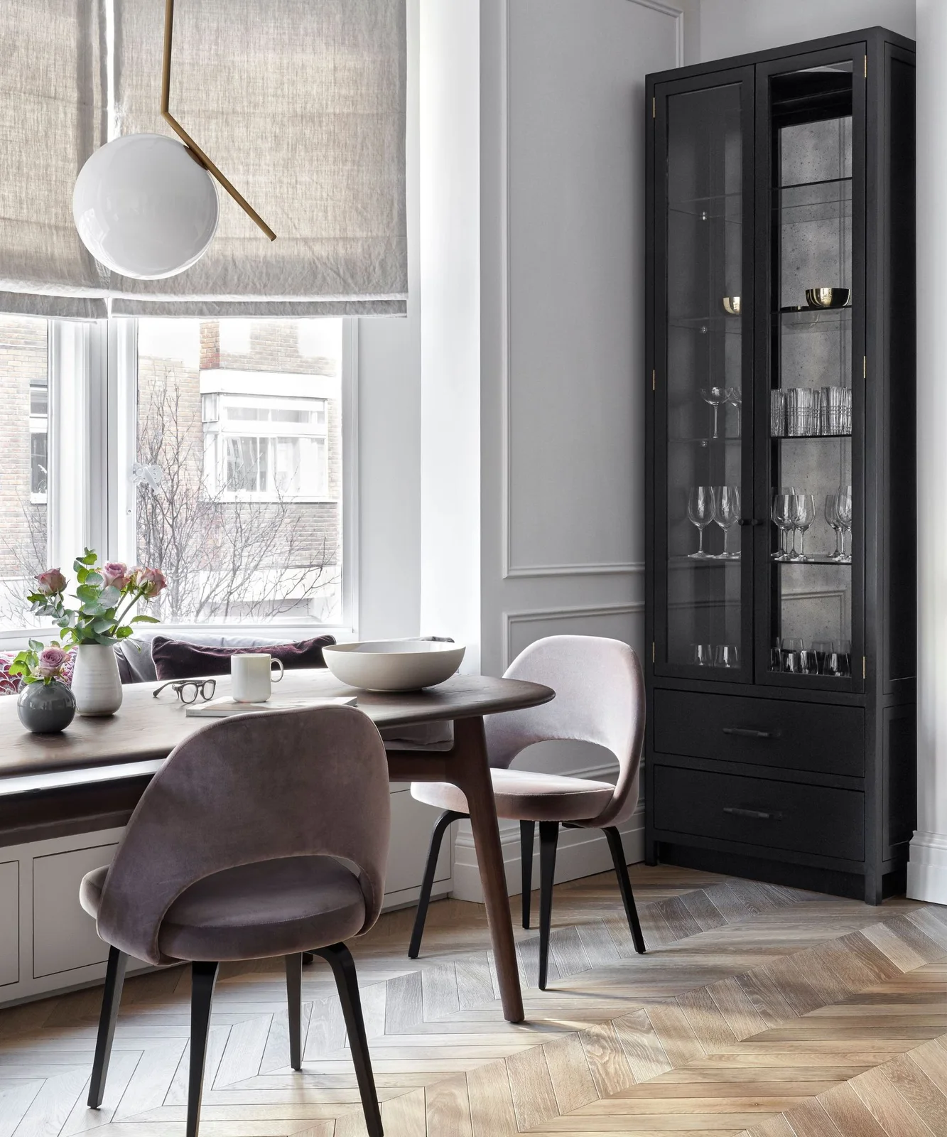 A breakfast table and pale upholstered chairs sit beneath a white globe pendant beside a bay window and tall dark glass-front cabinet
