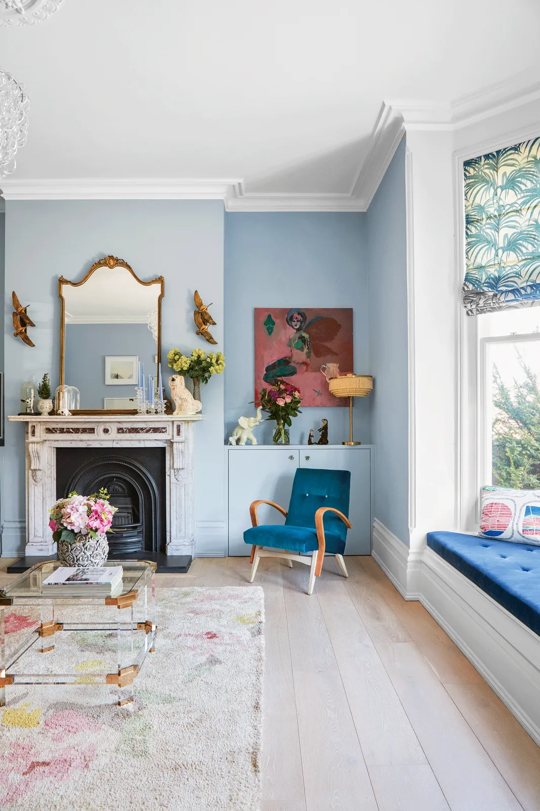 Pale blue walls surround a low window seat and colorful artwork beside a fireplace.