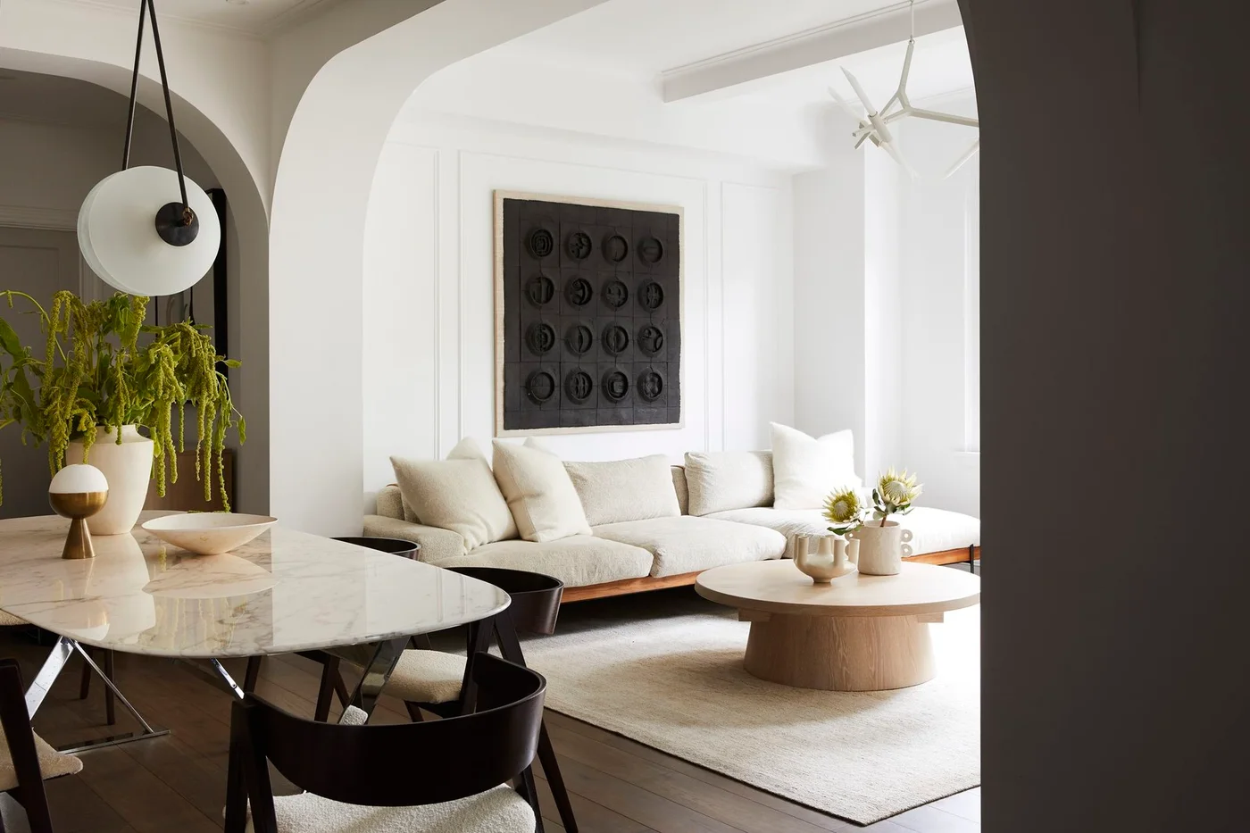 A pale sofa and round wood coffee table sit beyond a dining table beneath broad curved openings with a large dark artwork on the living-room wall