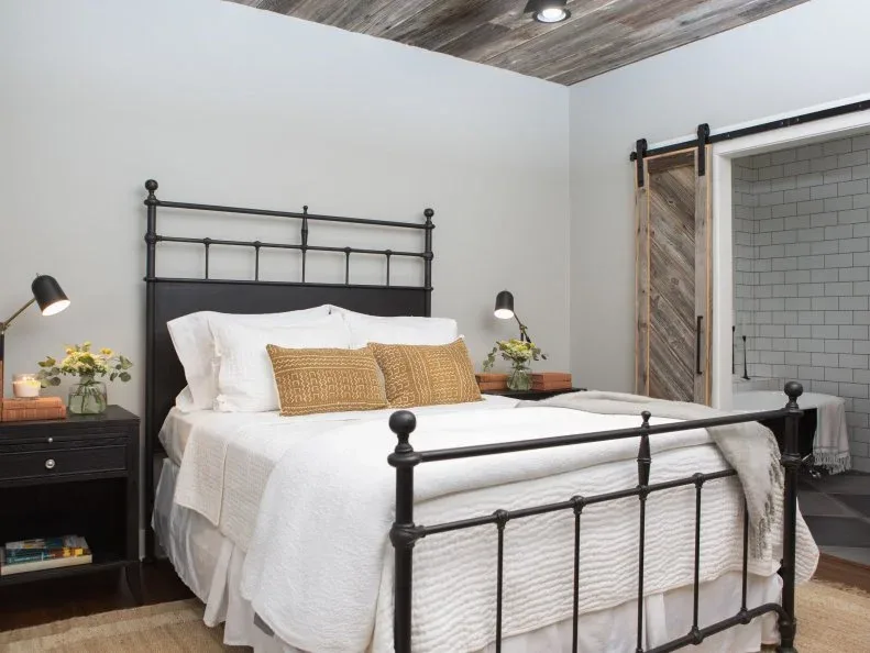 Textured wood bedroom ceiling above a dark metal bed, pale bedding, ochre cushions and a sliding wood door