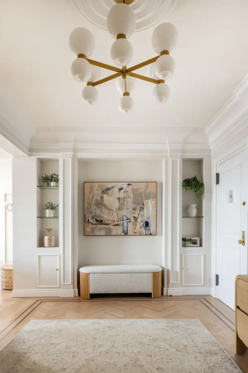 Gold-colored branching chandelier with white oval lights above pale paneled entry, padded bench, shelves and herringbone floor