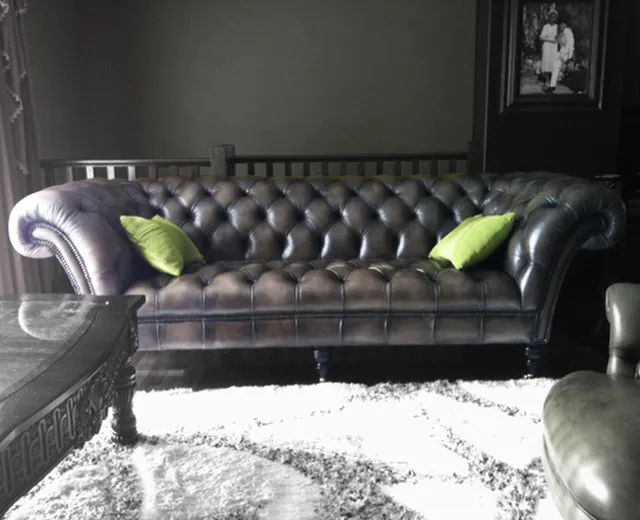 A black tufted leather sofa sits against dark walls with small green cushions and a pale rug.