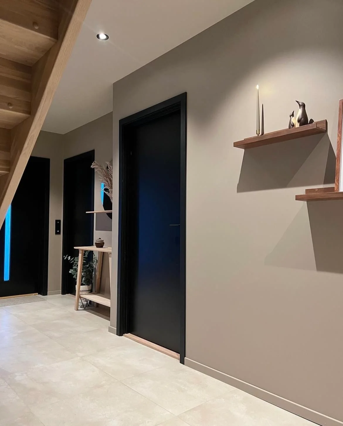 Wooden floating shelves stand beside dark doors and a wooden staircase against muted walls over a pale tiled floor