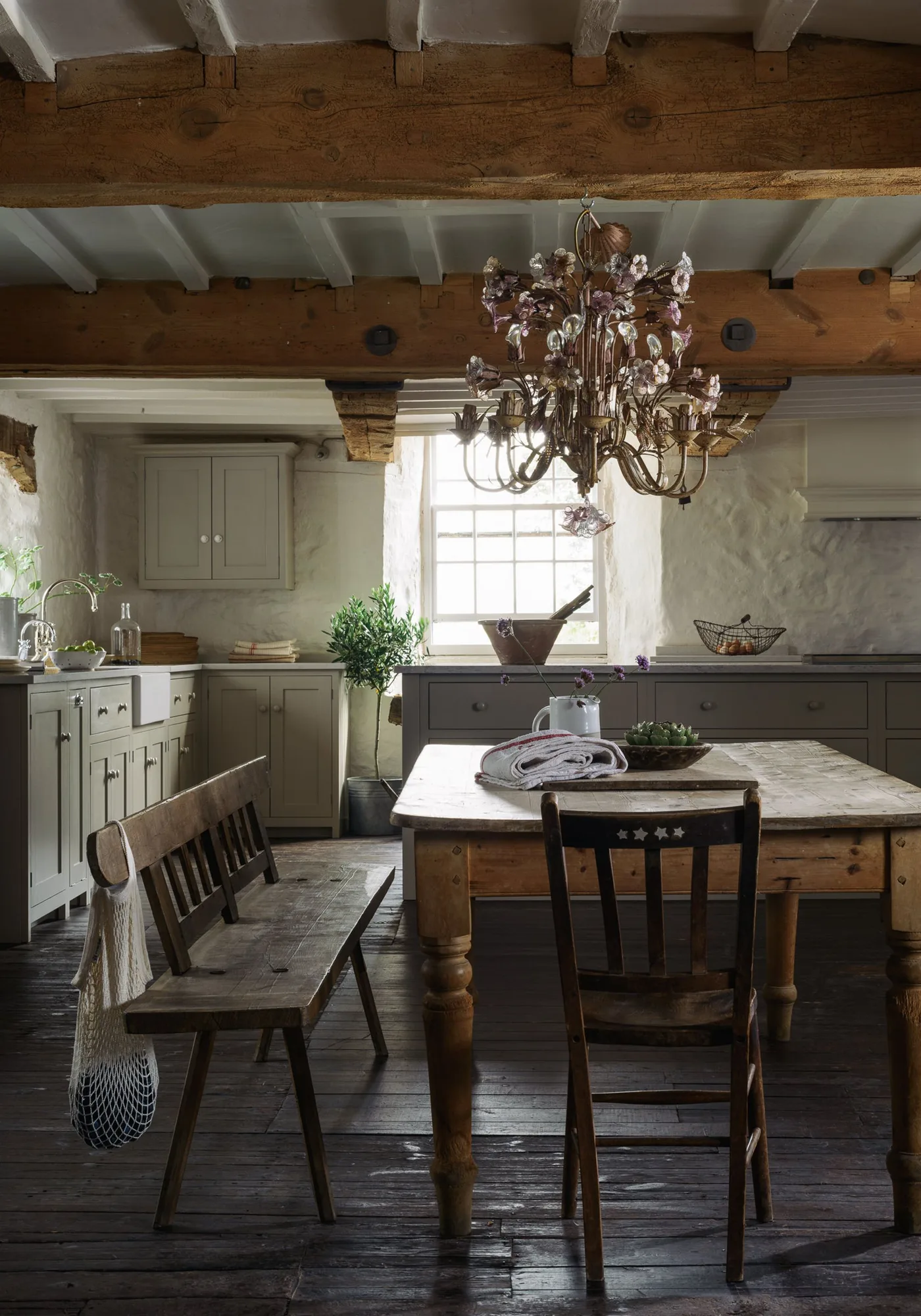 Cottage kitchen with gray-green cabinets, heavy wooden beams, wooden table and bench, dark floorboards and a decorative hanging light.