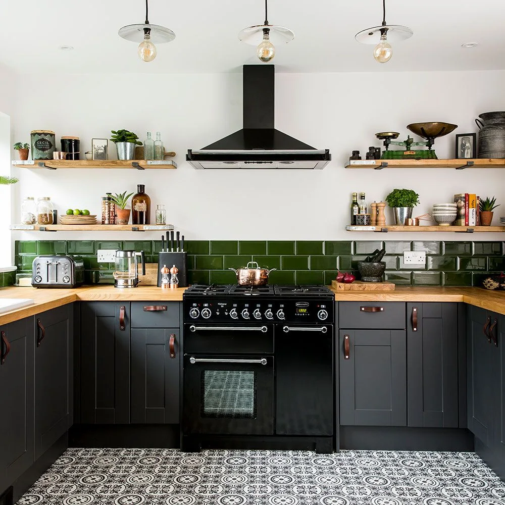 Charcoal gray lower cabinets with brown strap-style pulls sit beneath green rectangular tiles, wooden counters and open wooden shelves
