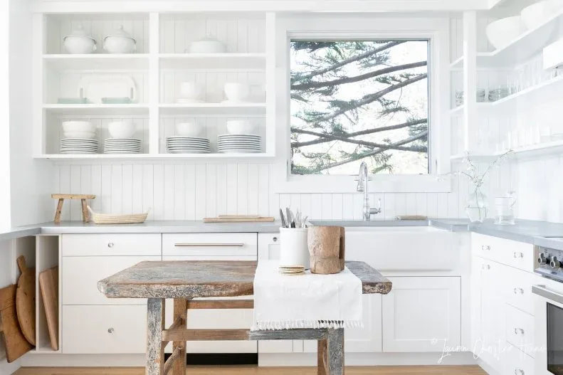 White kitchen with open shelves, paneled walls, small weathered wood table and simple stool near a bright sink window