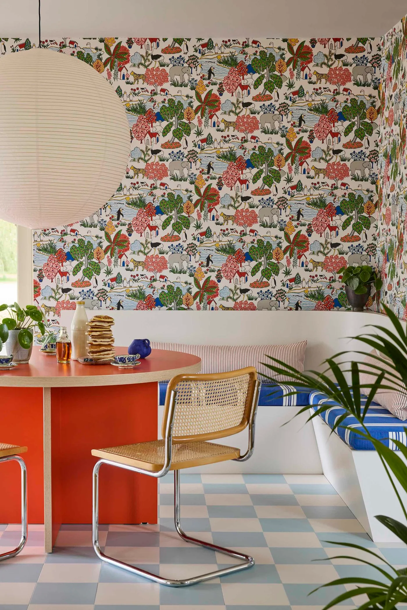 Orange round dining table beside a white corner bench, colorful wallpaper, woven-backed chairs and a large white pendant