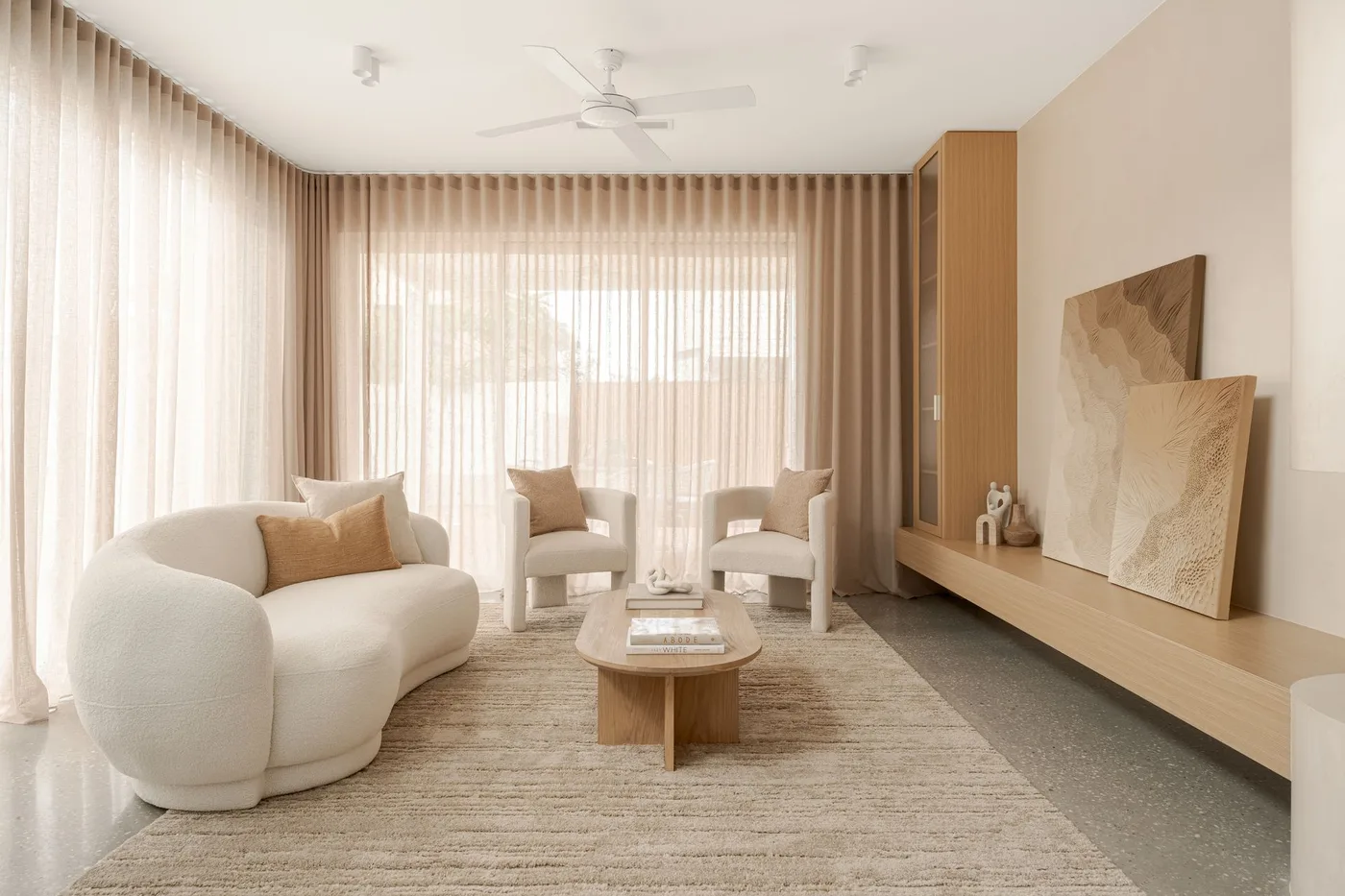 Pale curved living room seating beside a low wood ledge, tall wood storage unit and long light curtains