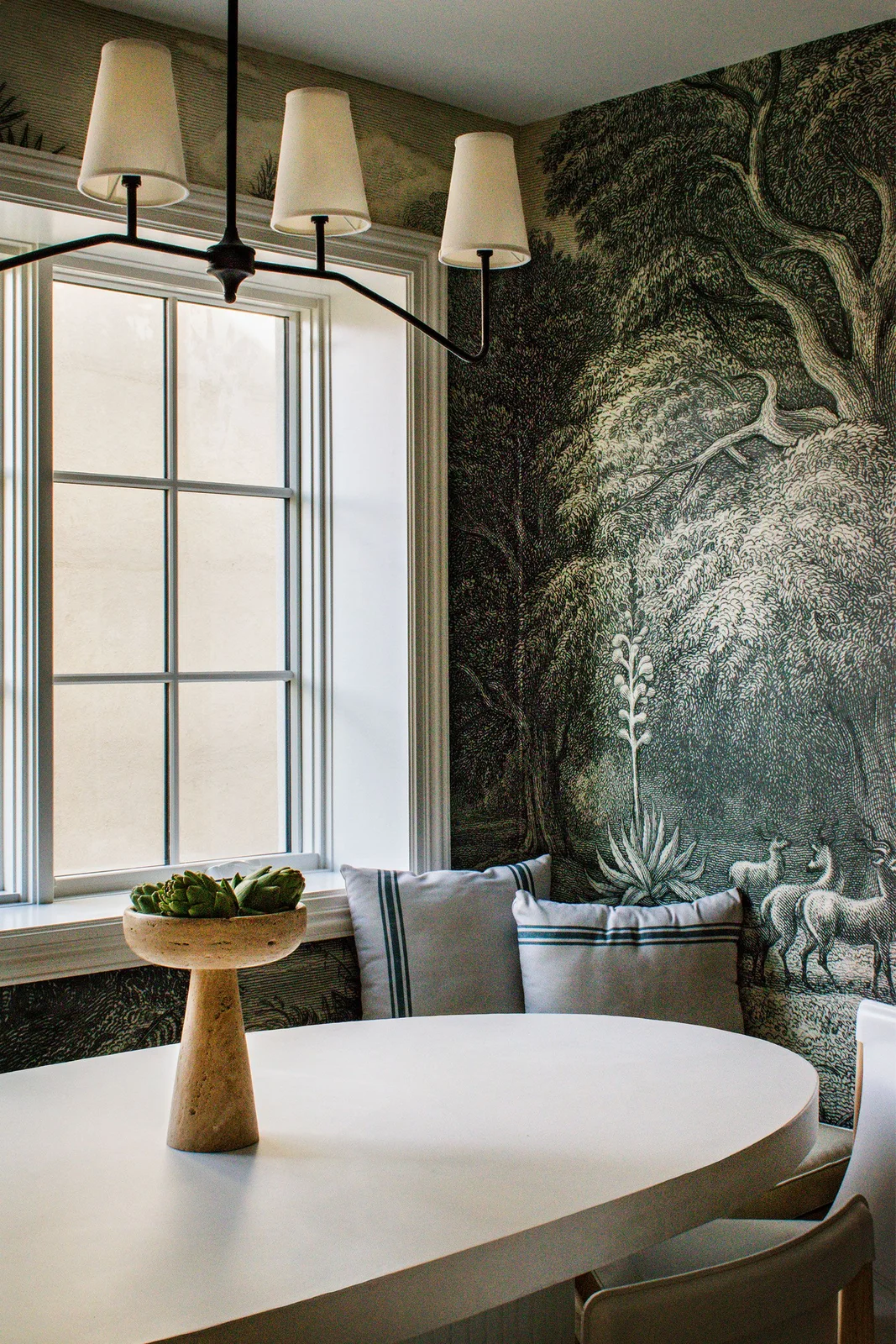A dark detailed woodland mural surrounds a pale oval dining table, gray striped cushions and a black multiarm light with pale tapered shades