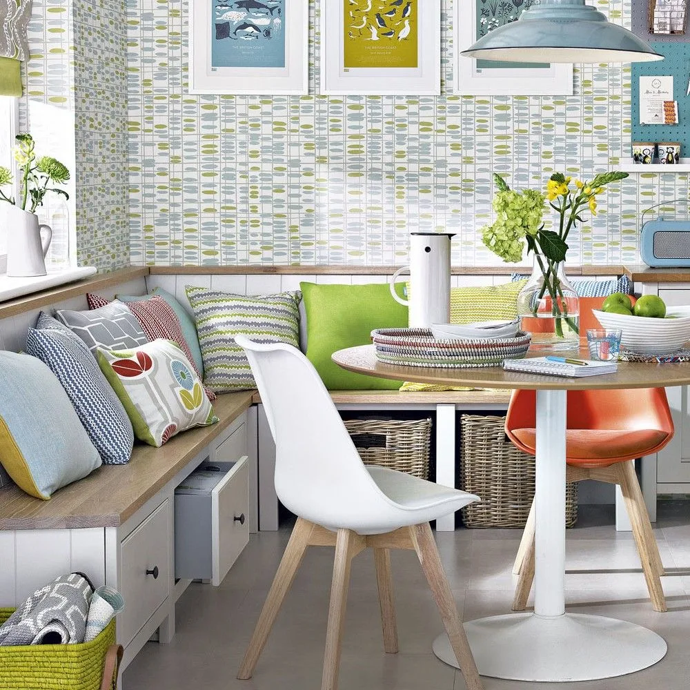 Fitted corner bench with drawers and open basket compartments, mixed cushions, patterned wallpaper, a white pedestal table, pale and orange chairs, and framed prints.