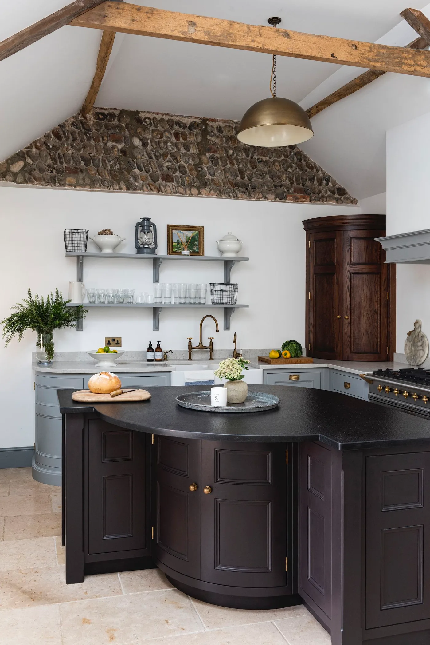 Cottage kitchen with a dark curved-front island, pale gray perimeter cabinets, exposed stone high on the wall, wooden beams and open shelves.