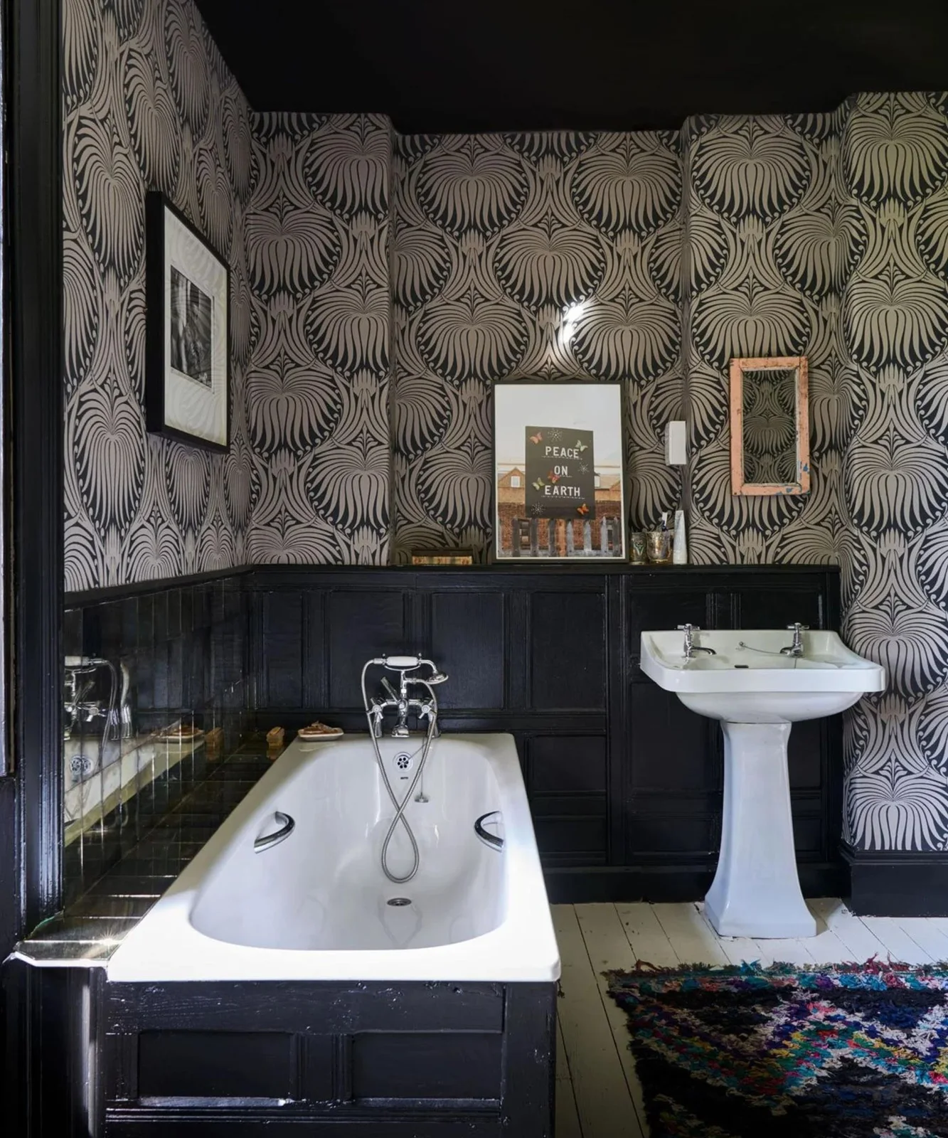 Black paneling surrounds a white bath and pedestal basin beneath dense monochrome wallpaper beside a multicolored rug
