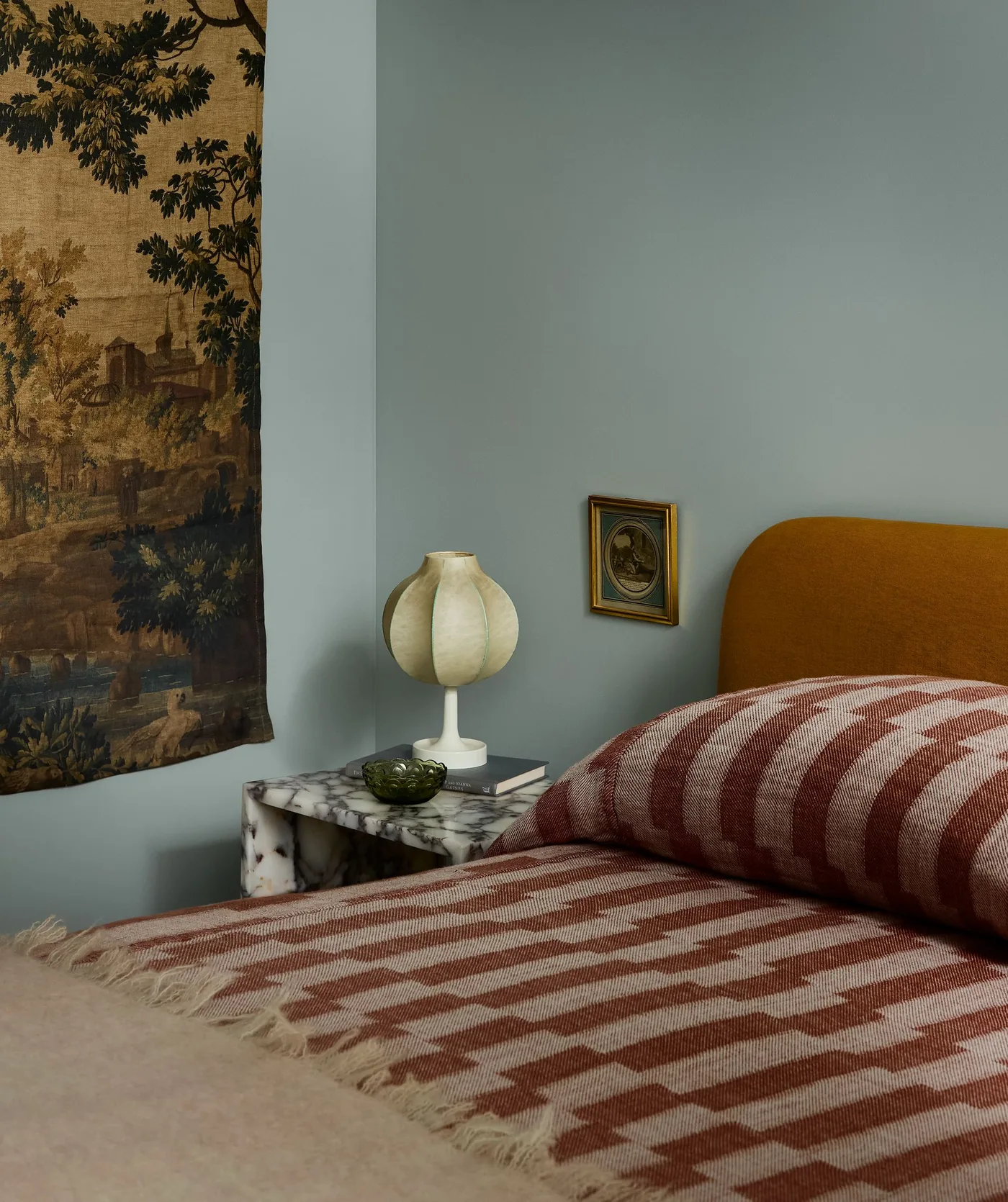 Bedroom with an ochre headboard, rust and white zigzag blanket, pale blue wall, landscape wall hanging and sculptural white bedside lamp