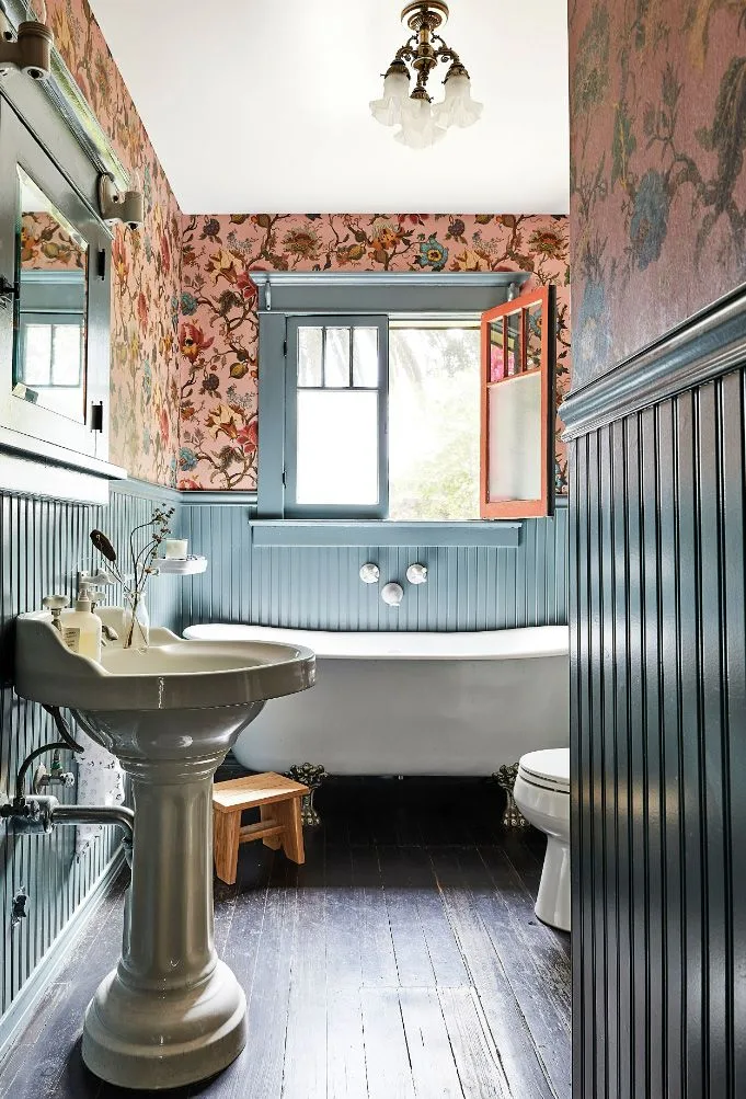 Narrow bathroom with deep blue vertical paneling, pink floral wallpaper, a pedestal sink, white bath and an open window.