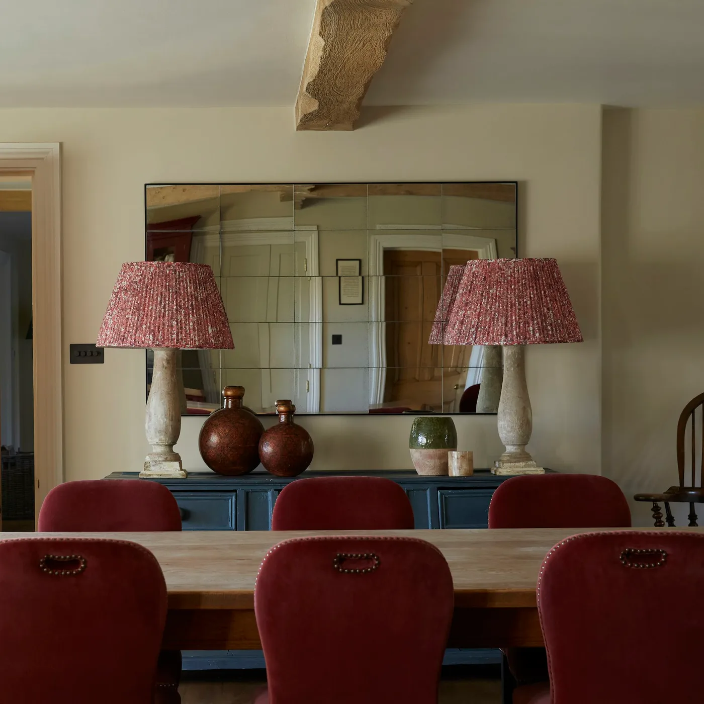 Wide rectangular mirror above a blue sideboard with two red-patterned lamps behind red dining chairs