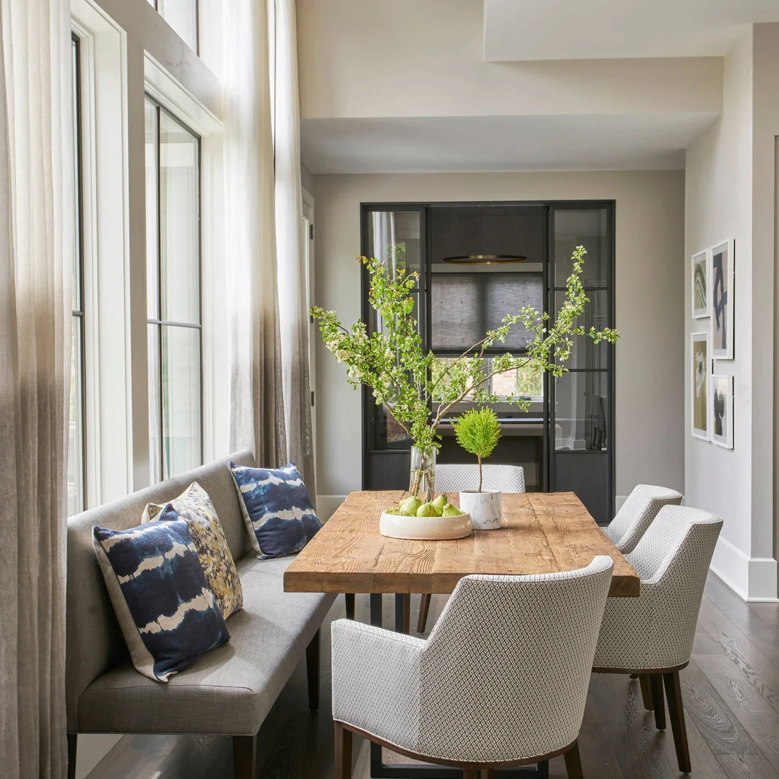 A wood rectangular dining table stands beside a gray upholstered window bench, pale chairs, blue patterned cushions and a dark-framed doorway