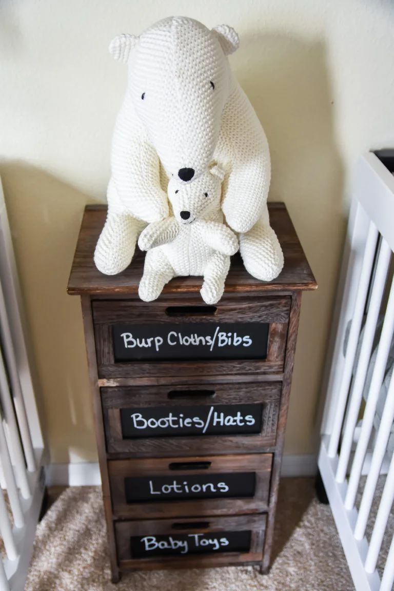 A narrow wood nursery drawer unit has dark labels for cloths, hats, lotions and toys with two pale bear ornaments on top beside white crib edges