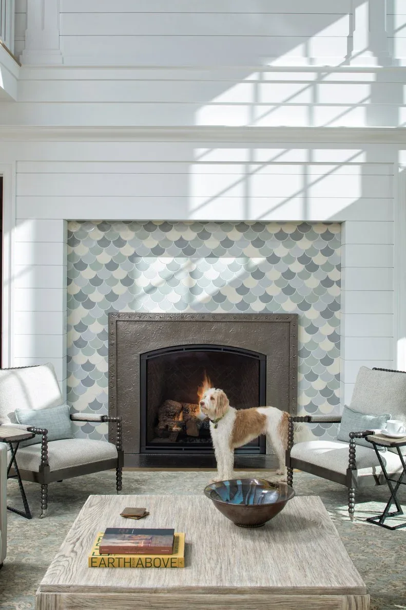 Pale scalloped fireplace tile below white horizontal boards with black arched opening and pale chairs