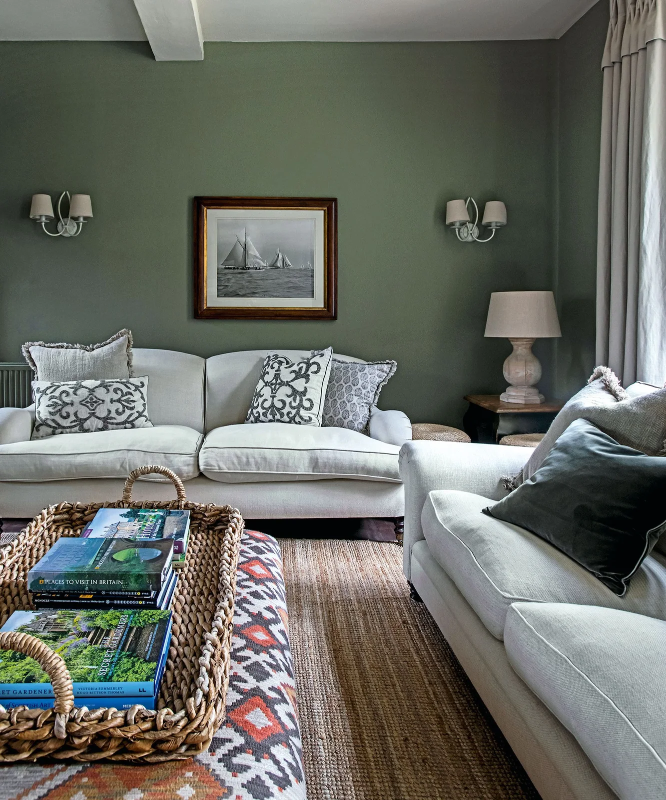 Pale sofas with patterned cushions face a patterned footstool and woven tray against olive green walls